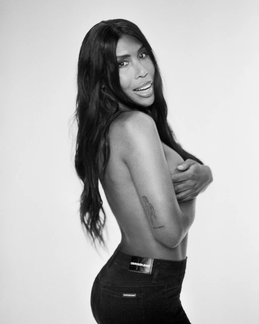 Black and white portrait of an adult woman with long dark hair, wearing jeans, covering her chest with her hand in a photo studio setting.
