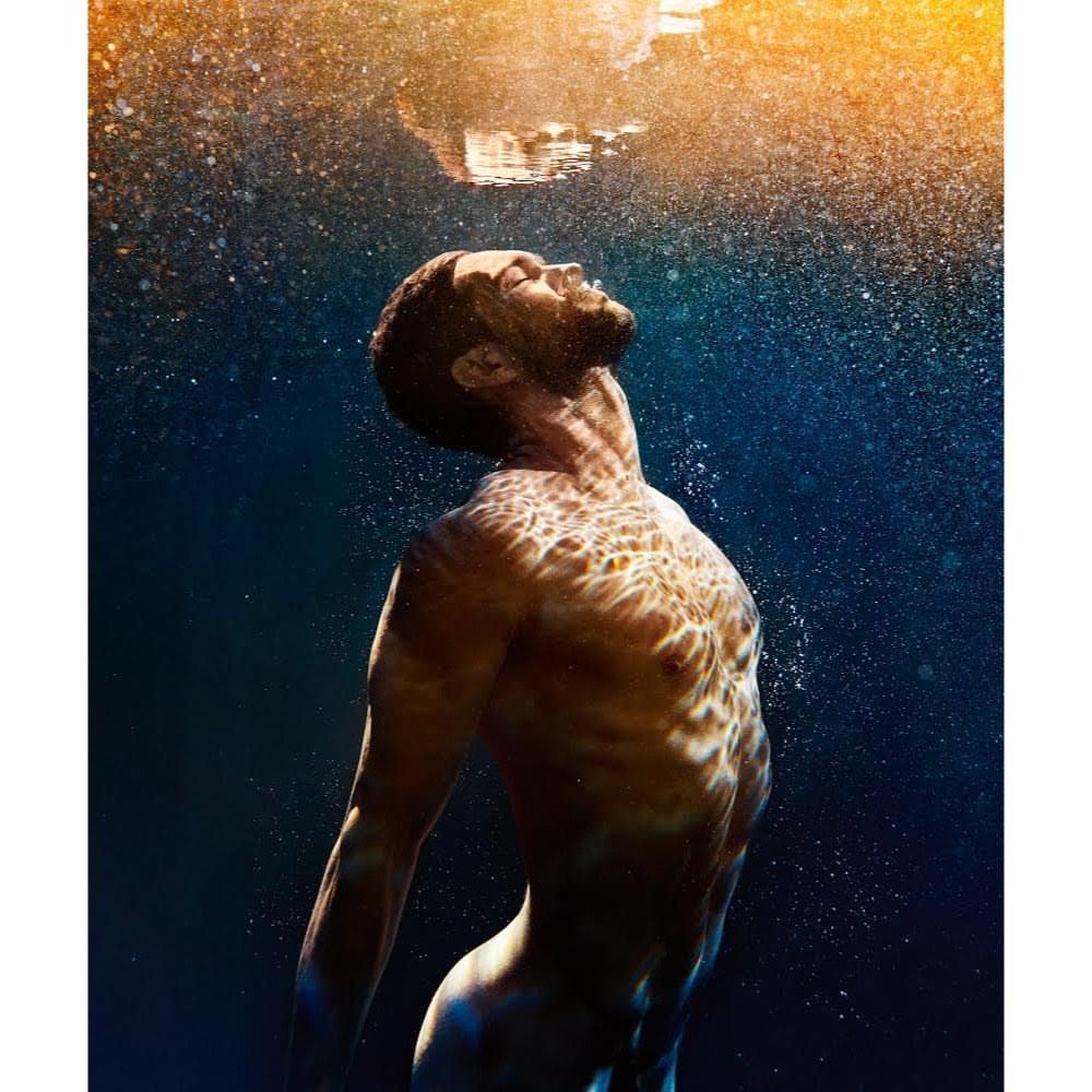 Underwater portrait of a fit man with light reflecting on his skin, eyes closed, against a dark blue background with golden light at the top.
