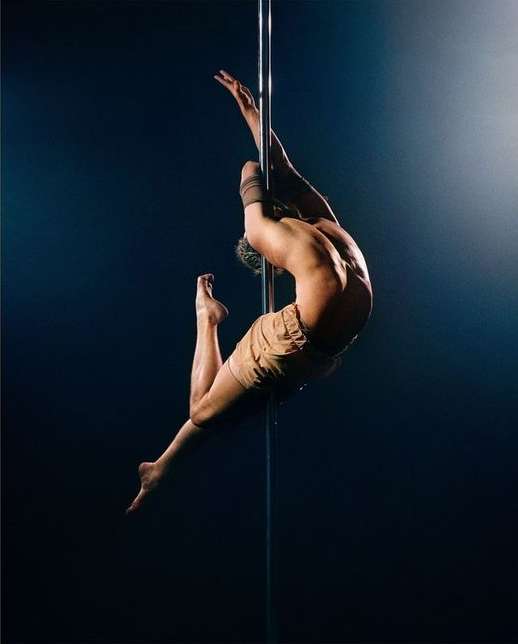 A muscular man is performing pole dancing, showcasing strength and control with a dynamic pose against a dark backdrop with a light source.