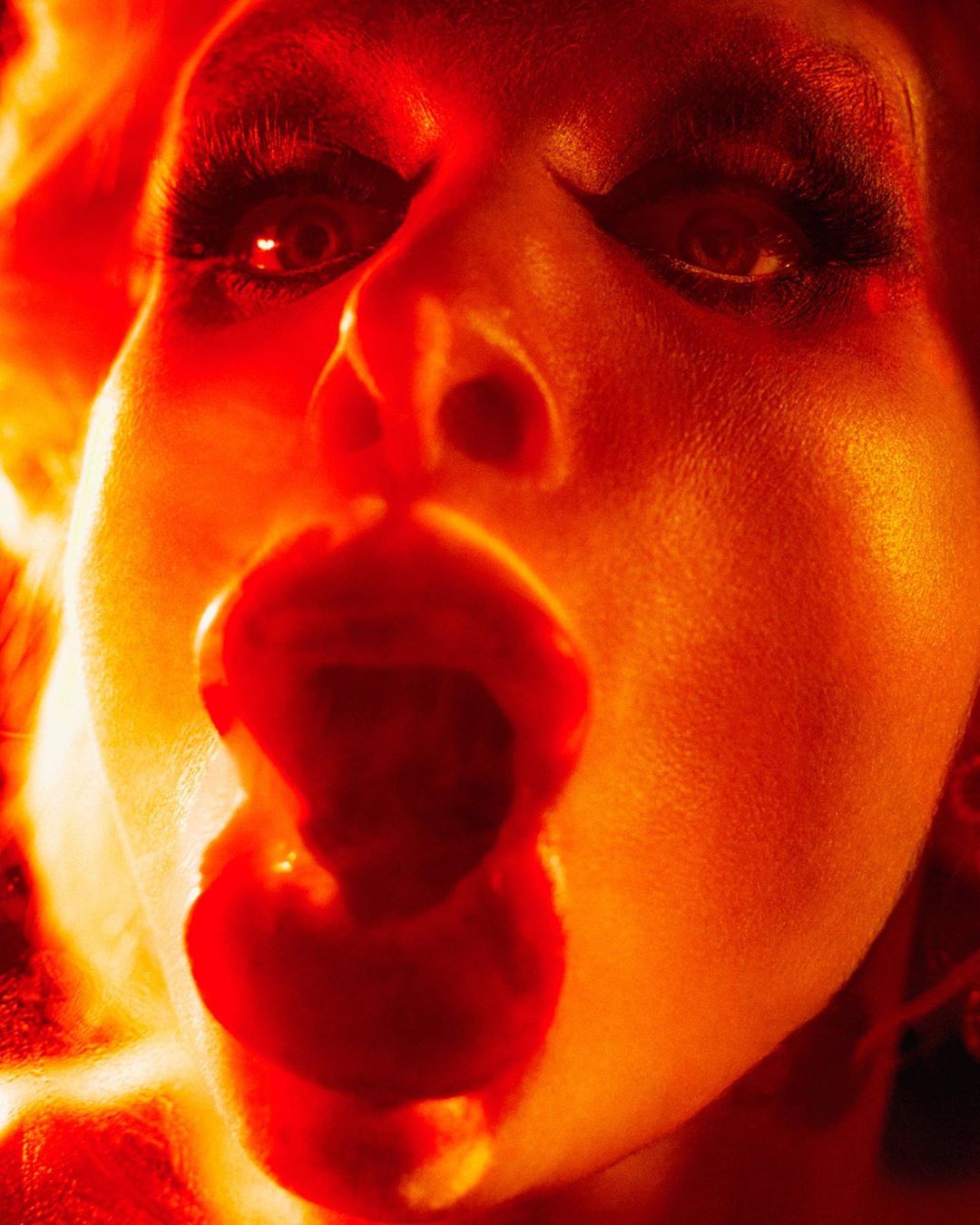 Close-up portrait of an adult female with dramatic make-up and orange-toned lighting with mouth open.