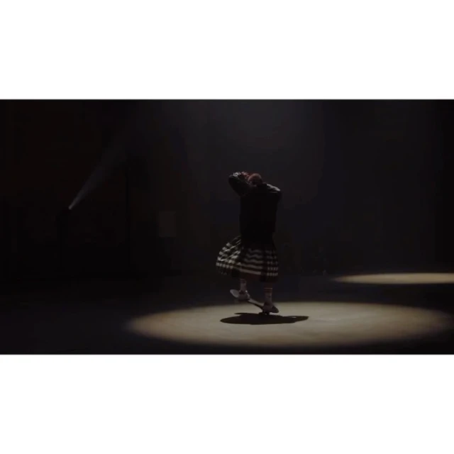 A dancer in a black jacket and patterned kilt performs in a spotlight against a dark background, showcasing movement and theatrical lighting.