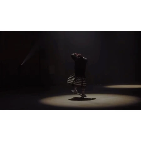 A dancer in a black jacket and patterned kilt performs in a spotlight against a dark background, showcasing movement and theatrical lighting.