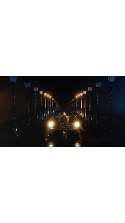 A man stands in front of a classic black car with headlights on, on a dark city street lined with vintage streetlights.