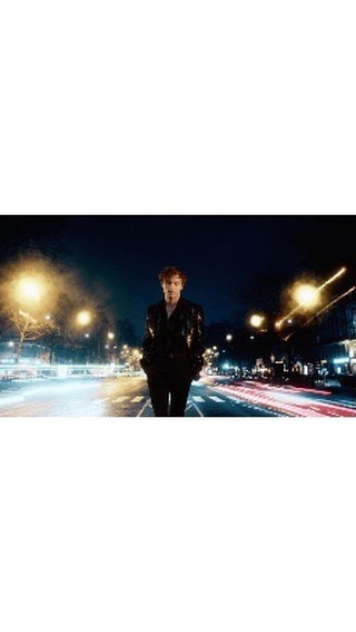 An adult male with red hair stands on a city street with buildings and blurred light trails in the background at dusk or nighttime.