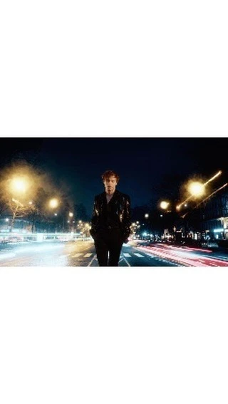 An adult male with red hair stands on a city street with buildings and blurred light trails in the background at dusk or nighttime.