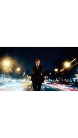 An adult male with red hair stands on a city street with buildings and blurred light trails in the background at dusk or nighttime.