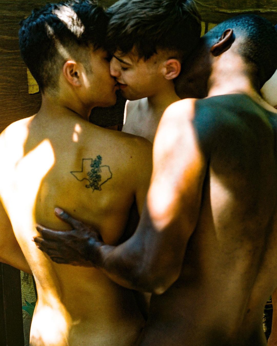 Intimate gathering featuring three shirtless adult males, with one fair-skinned man and another kissing, in a sunlit outdoor setting.