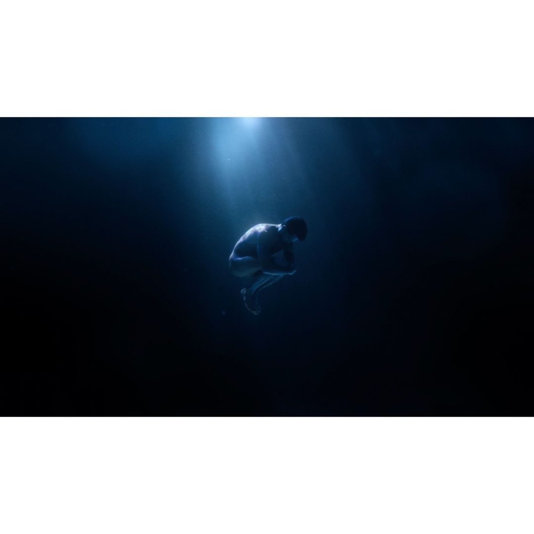 Adult male in fetal position underwater with bright light shining down on him, creating a mysterious and ethereal scene.