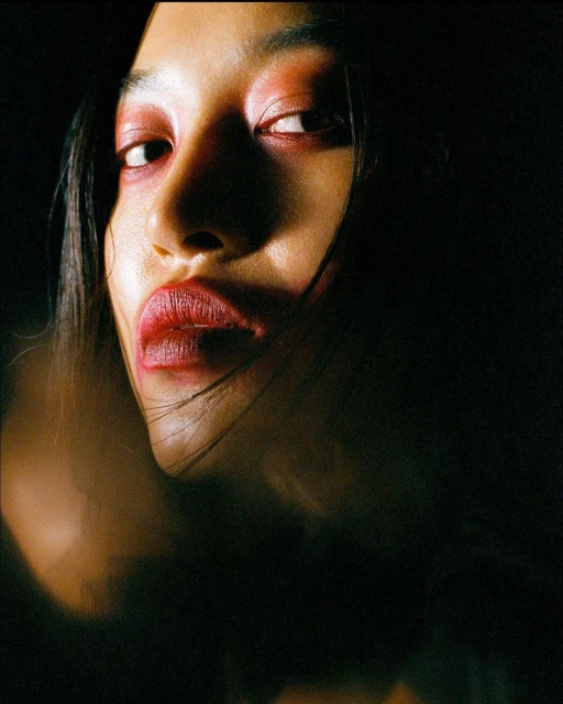 Close-up portrait of an adult female with red makeup on her lips and eyelids, set against a dark and moody background with dramatic lighting.