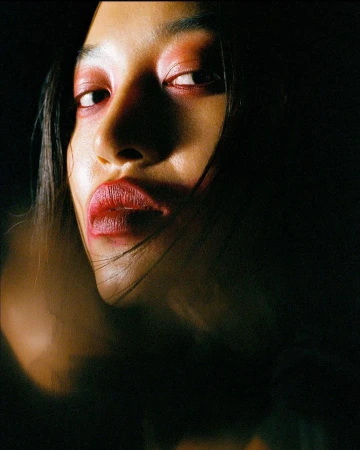 Close-up portrait of an adult female with red makeup on her lips and eyelids, set against a dark and moody background with dramatic lighting.