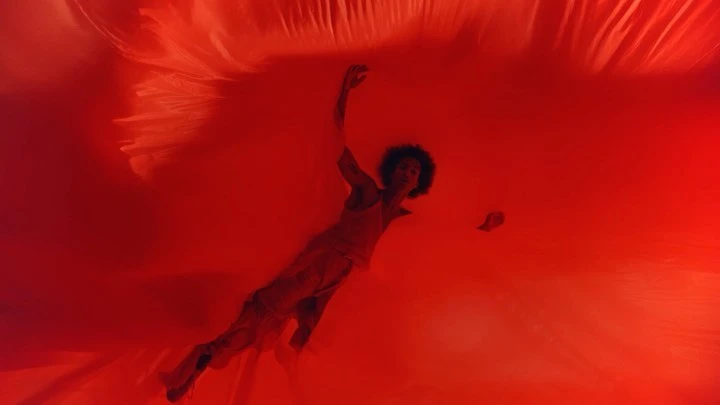 Adult female in a red tank top and pants submerged in red fabric. She has curly dark hair and her arm is raised above her head.