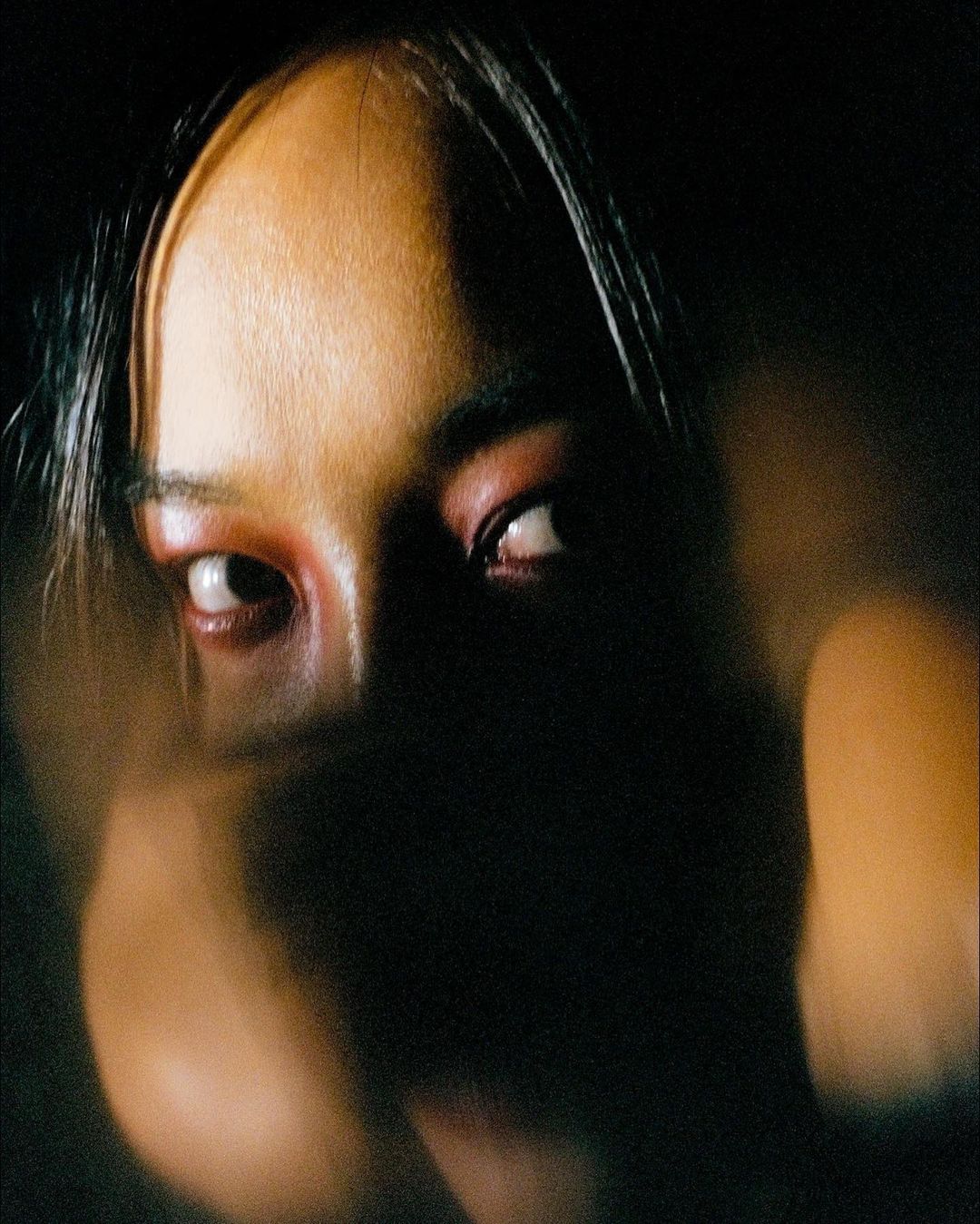 Close-up portrait of an East Asian adult female with red eye shadow in a dark indoor environment, creating a dramatic visual effect.