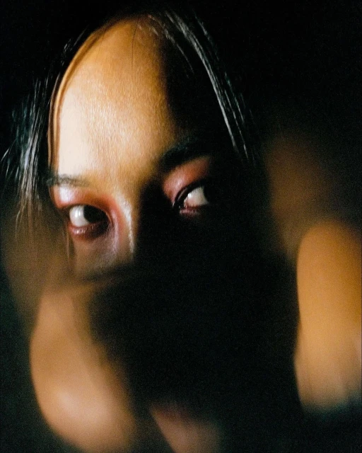Close-up portrait of an East Asian adult female with red eye shadow in a dark indoor environment, creating a dramatic visual effect.