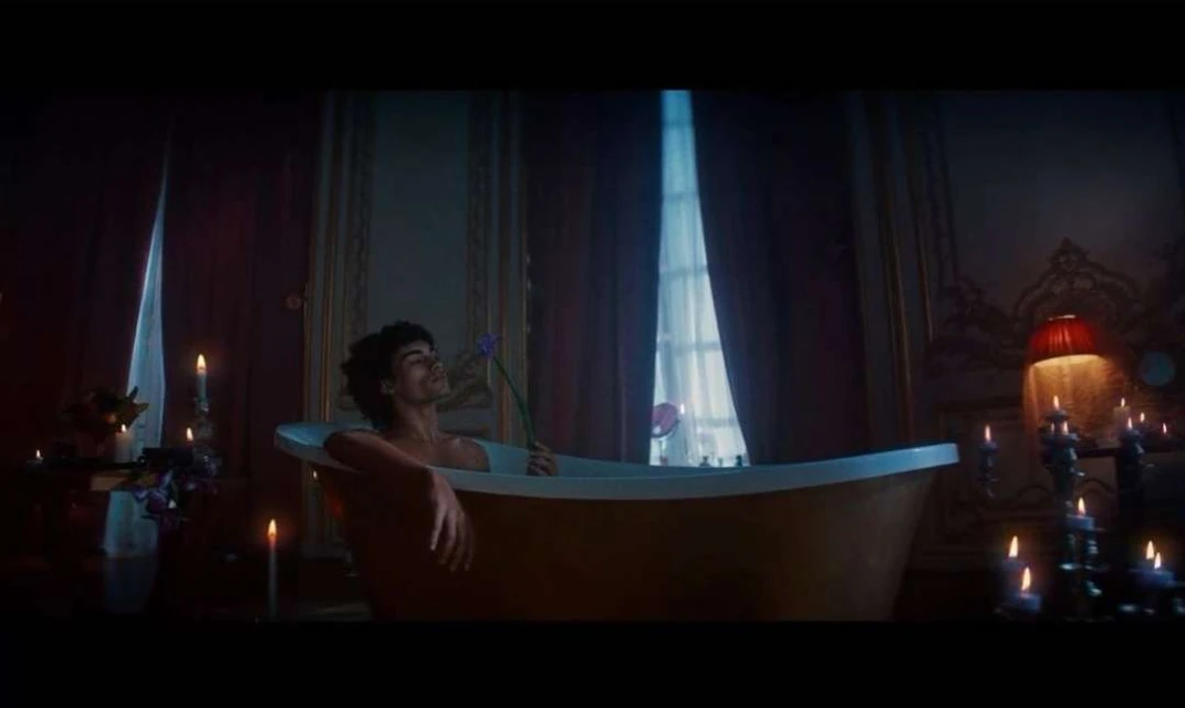 Man relaxing in a bathtub with a flower, surrounded by candles in an ornate bathroom with dark red walls and white window curtains.