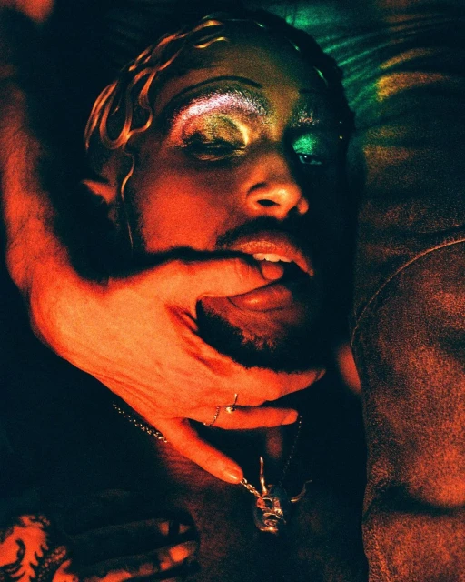 Adult male portrait with stylized makeup, fair skin, hand in mouth, jewelry, set indoors with warm lighting and orange and green tones.