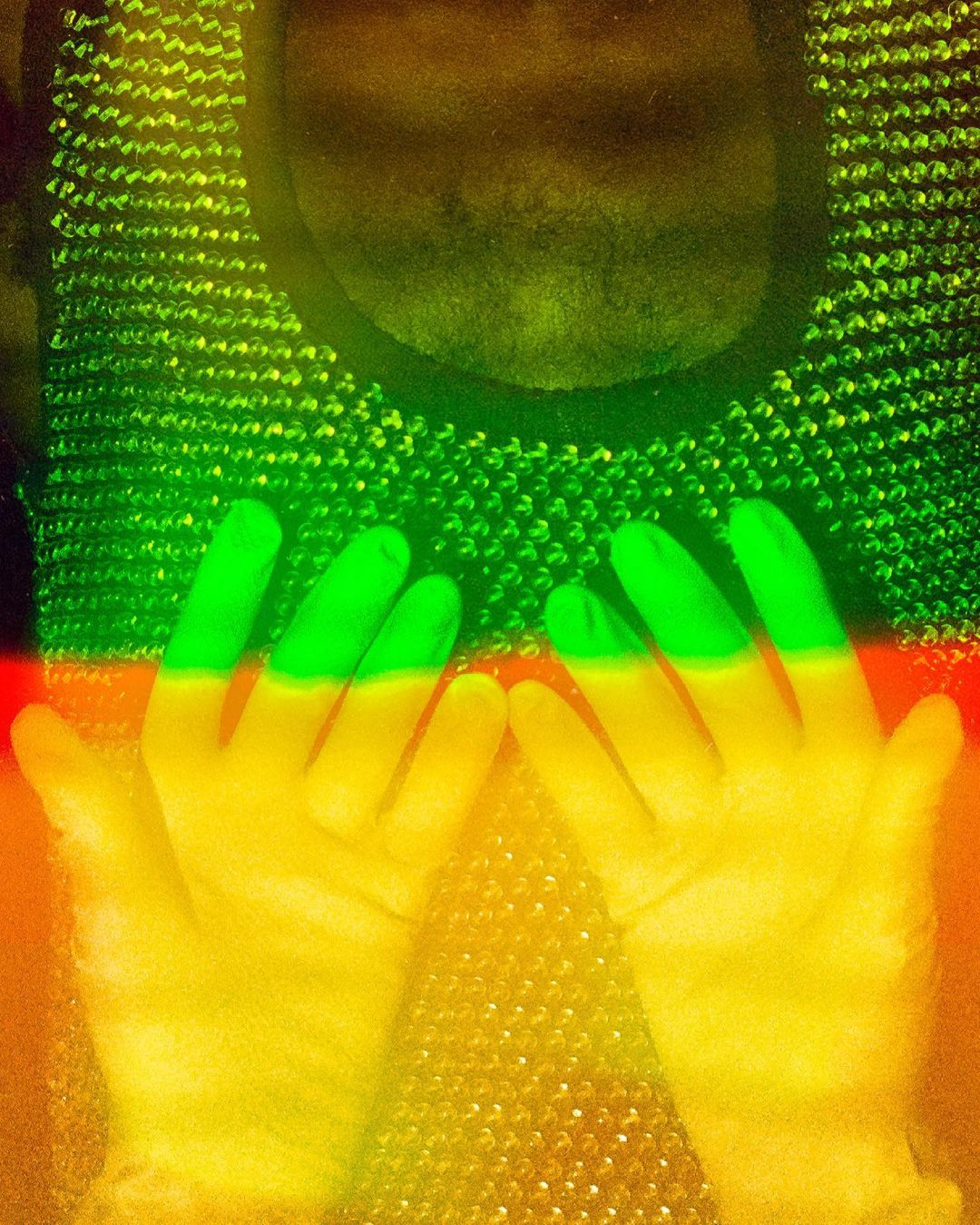 Abstract studio shot of a person's hands and upper torso wearing chainmail, with strong yellow, green, and red color grading.