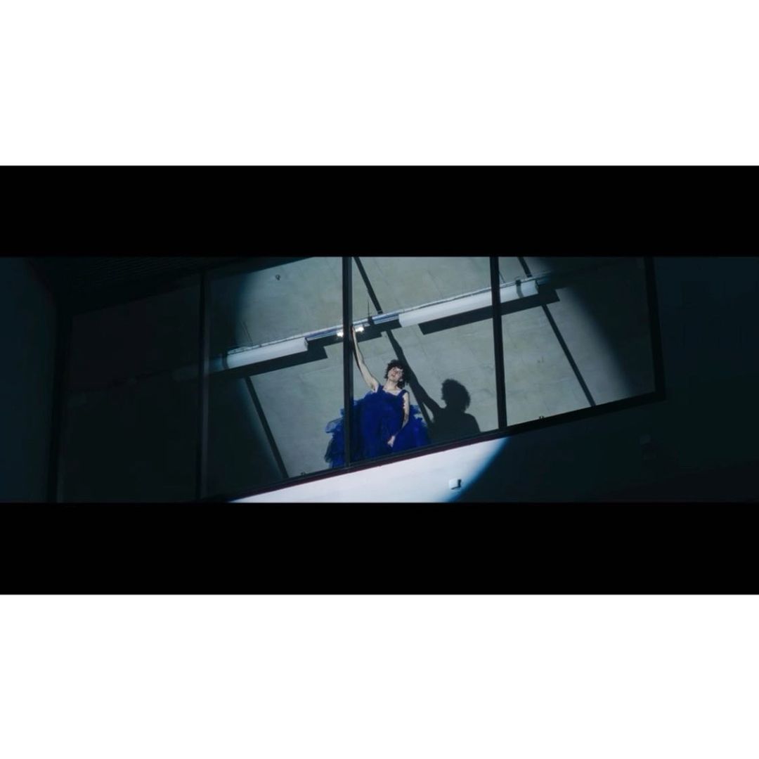 Adult woman in a blue gown is viewed through a window with geometric grids, shadowed, lit by a spotlight in a dark interior space.
