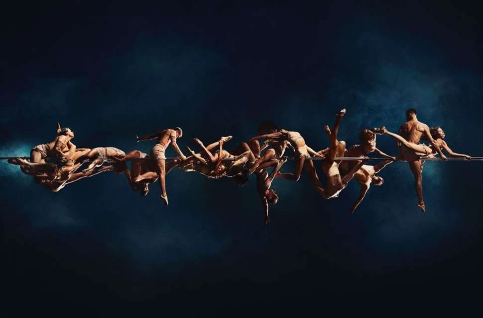 A group of dancers performing on a suspended bar against a dark blue background, showcasing a range of acrobatic poses.