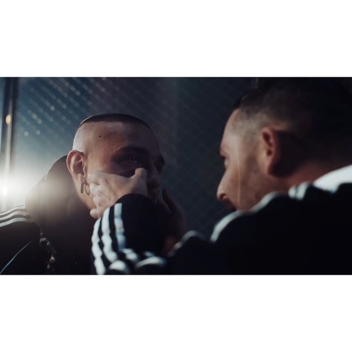 Man looks into a mirror, face partially obscured by his hands, wearing an Adidas tracksuit jacket with the three-stripe design on the sleeves.