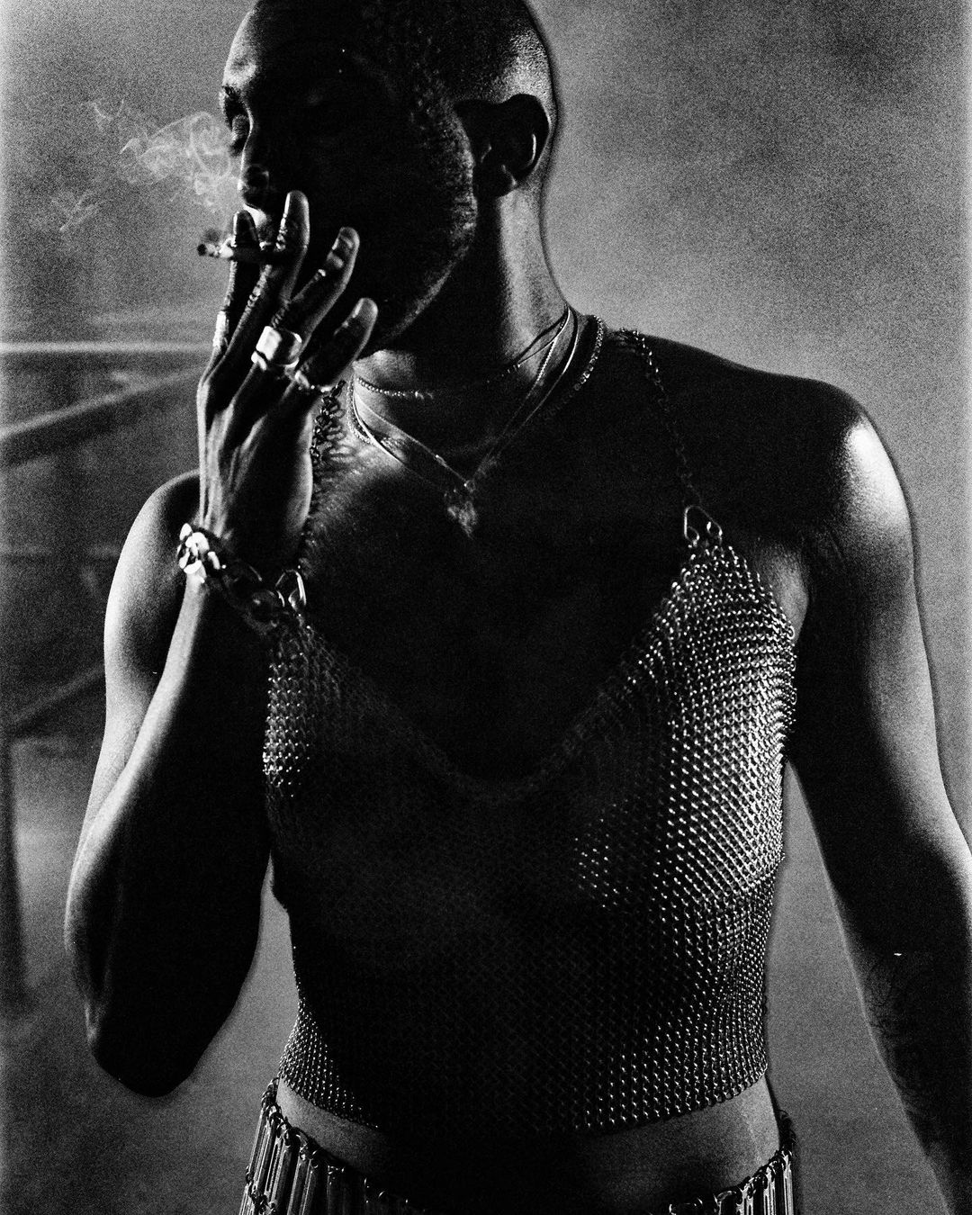 Black and white photo of a Black male with a shaved head smoking a cigarette and wearing a metal chainmail top with jewelry.