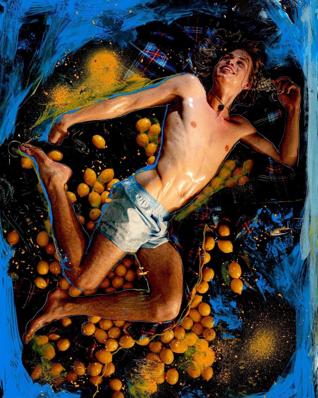 Artistic portrait of a fair-skinned adult male lying on a bed of lemons against a black and blue painted background with expressive splashes.
