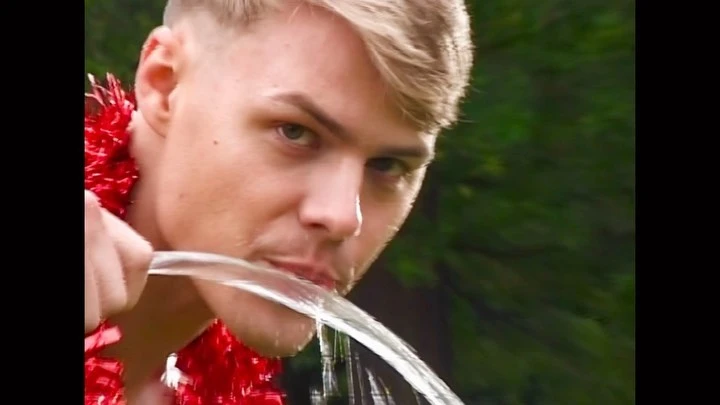 Close-up of a blond man wearing a red lei, pouring a stream of water into his mouth outdoors, with blurred green foliage background.