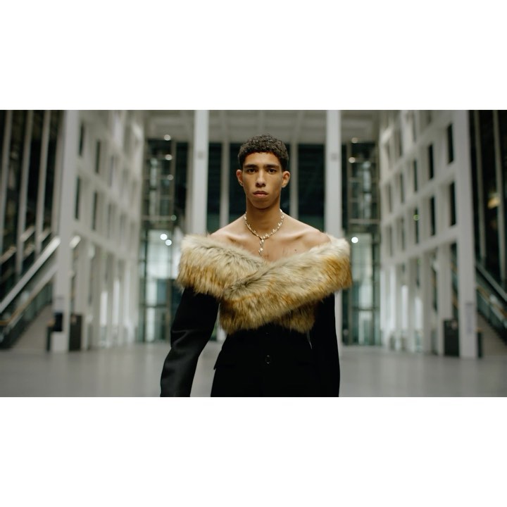 Portrait of a young person with short curly hair, wearing a fur stole and black suit in a building with white columns.