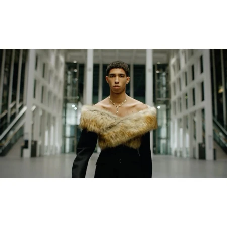 Portrait of a young person with short curly hair, wearing a fur stole and black suit in a building with white columns.