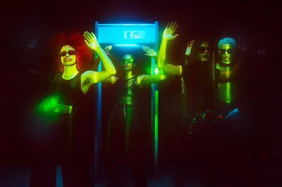 Group of four people standing with raised hands under a metal detector-like structure with blue and green lighting in a dark room.