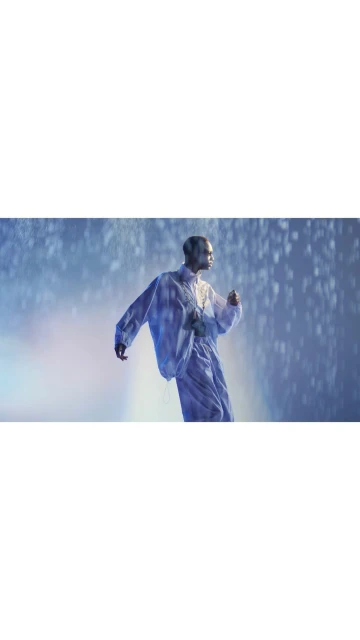 An adult male dances in a blue-toned indoor environment with rain effects and cool lighting, wearing a translucent long-sleeved top.