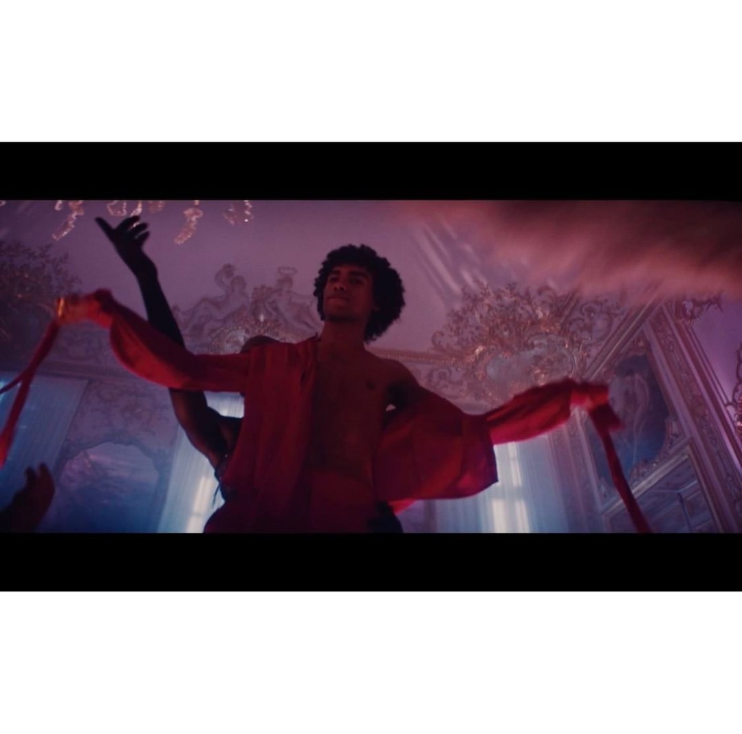 A man dances in a red robe in an ornately decorated room illuminated by pink and blue lighting in a theatrical performance.