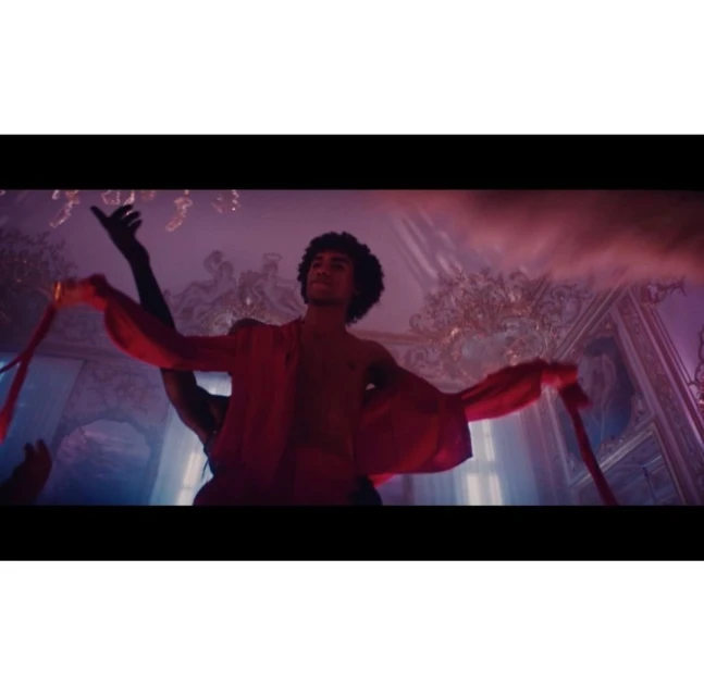 A man dances in a red robe in an ornately decorated room illuminated by pink and blue lighting in a theatrical performance.