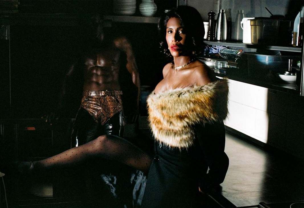 Woman in elegant dress with fur collar poses next to a muscular man with abs in a dark kitchen setting, highlighted by bright light.