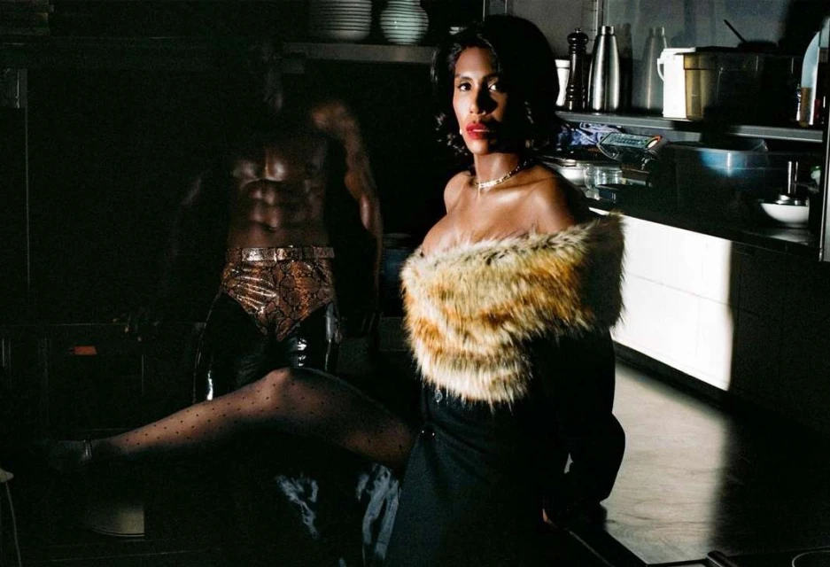 Woman in elegant dress with fur collar poses next to a muscular man with abs in a dark kitchen setting, highlighted by bright light.