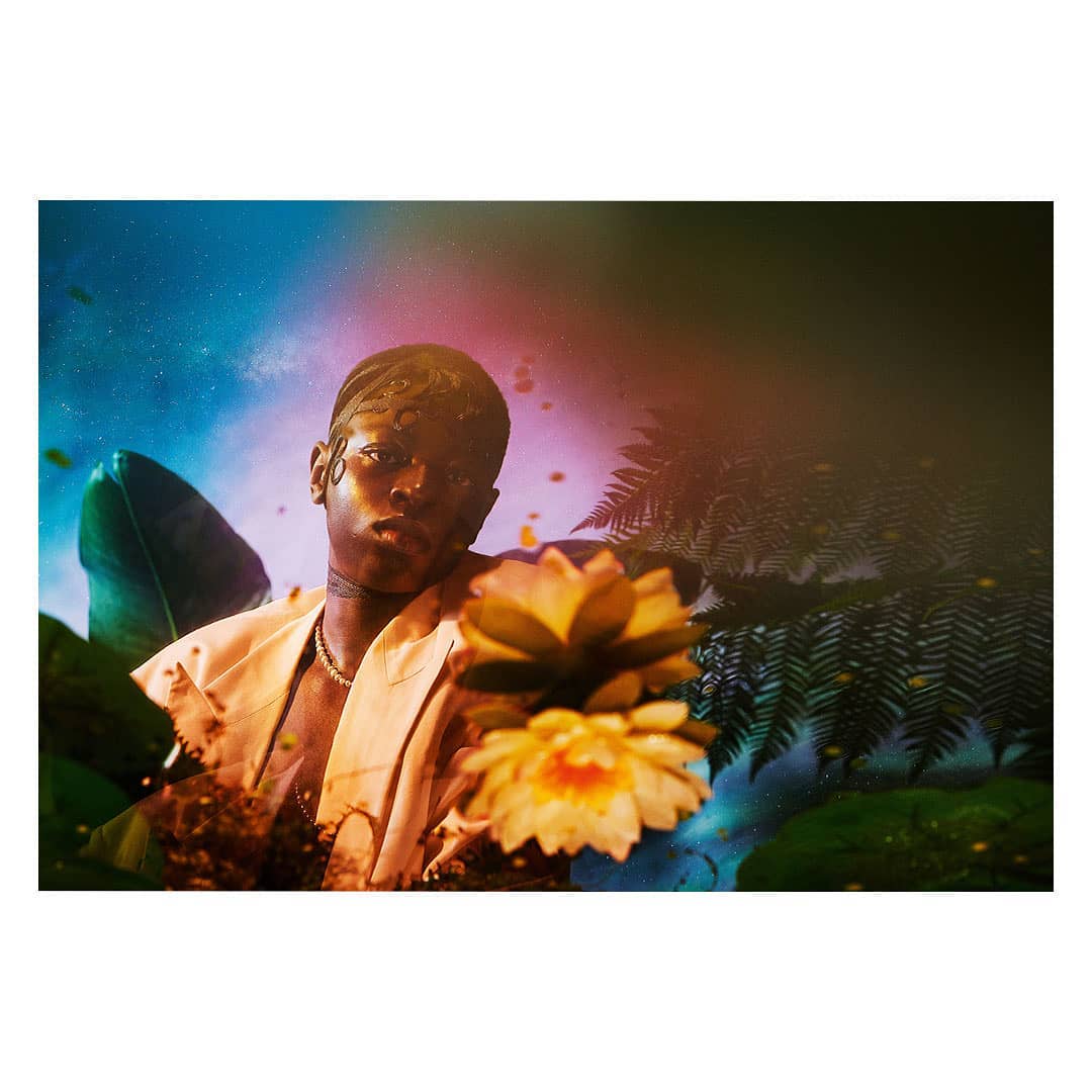 Adult male surrounded by tropical plants and flowers in surreal setting with blue and red sky, wearing beige suit jacket and beaded necklace.