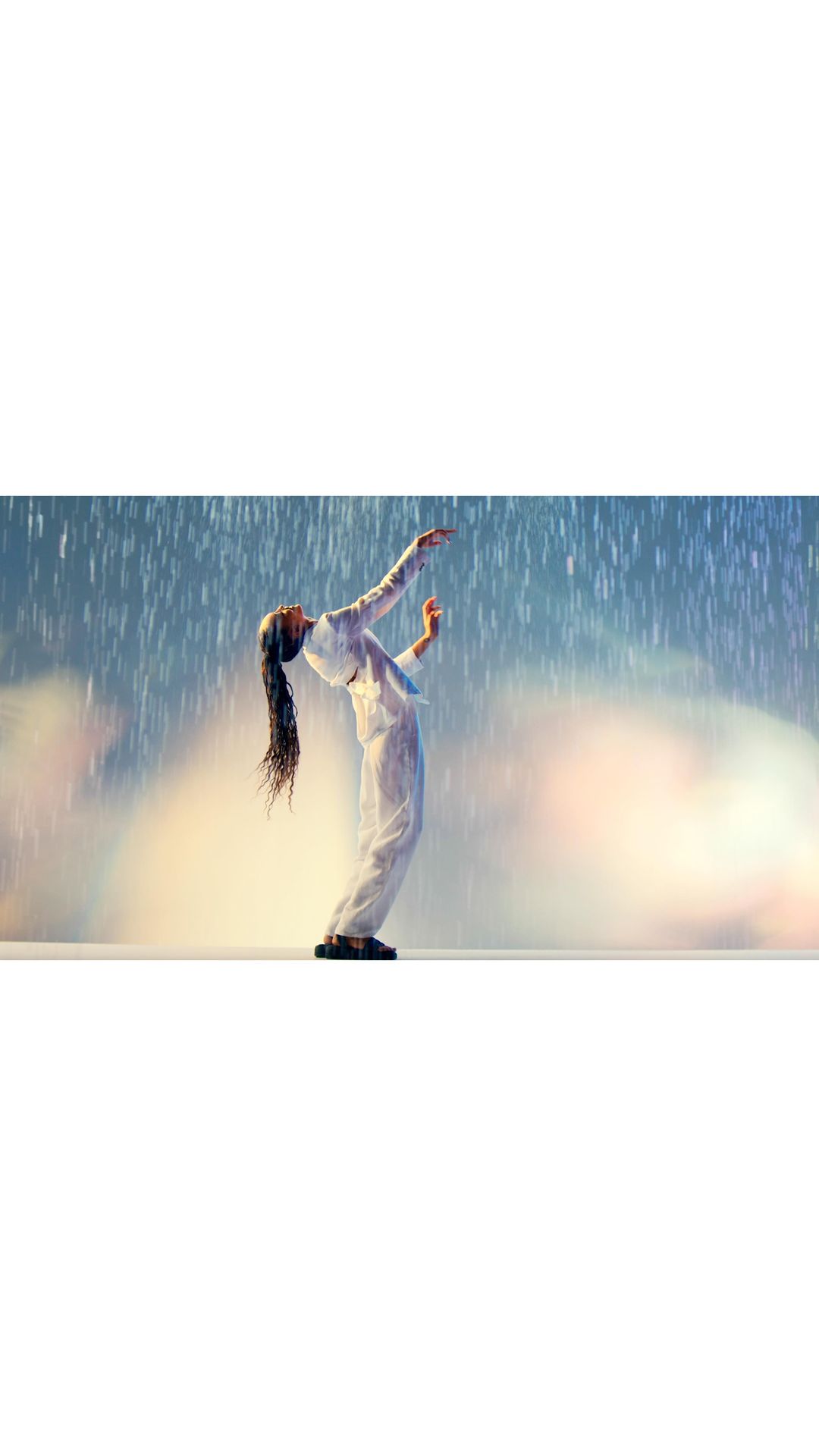 A woman in white arched backward with arms raised in a bright, artificial rain environment on a cream-colored stage.