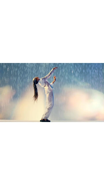 A woman in white arched backward with arms raised in a bright, artificial rain environment on a cream-colored stage.