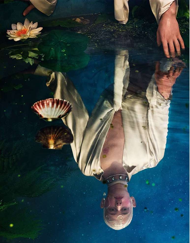 Person reflected in water with lily pads and scallop shells, wearing a white outfit and jewelry, creating a surreal and artistic scene.