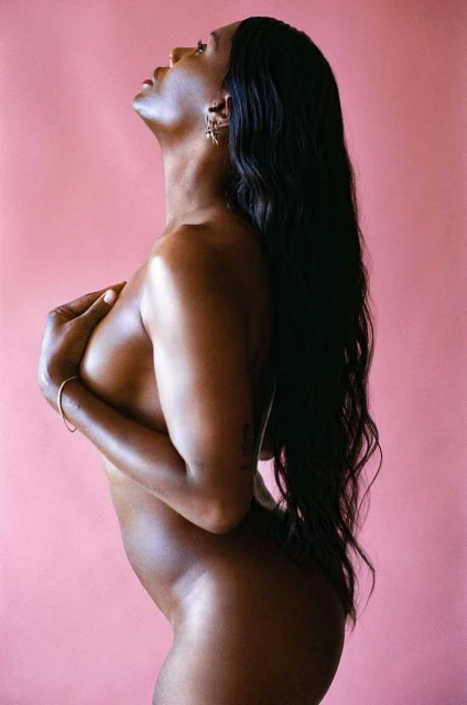Profile of an African American woman with long black hair, hands covering her breasts, posing against a pink background in a studio setting.