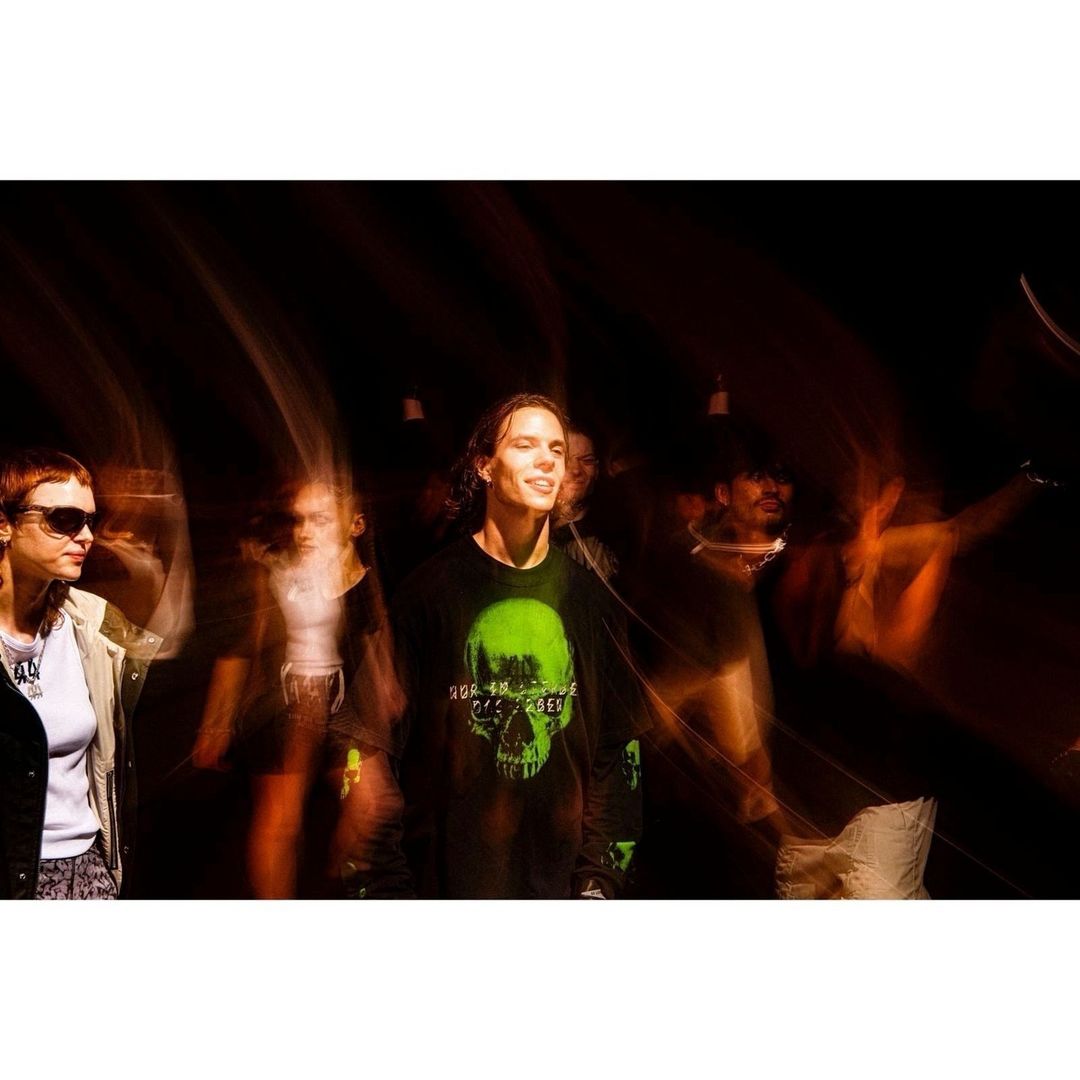 Group of people in an indoor setting, illuminated by bright lighting, creating motion blur. They are wearing casual clothing with a graphic tee.