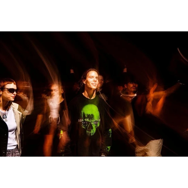 Group of people in an indoor setting, illuminated by bright lighting, creating motion blur. They are wearing casual clothing with a graphic tee.