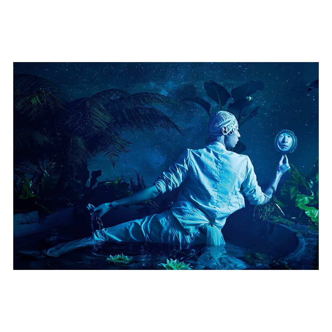 Person in white outfit, head covering, and bare feet sits near water, holding a small mirror against a backdrop of lush green plants and a starry sky.