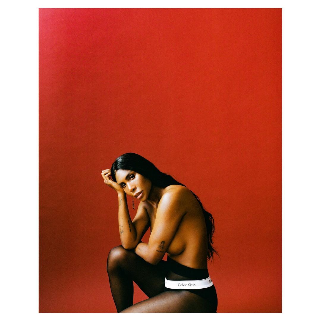 Adult woman with long black hair poses seated against a solid red background. She is wearing black leggings and Calvin Klein underwear.