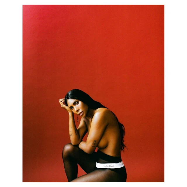 Adult woman with long black hair poses seated against a solid red background. She is wearing black leggings and Calvin Klein underwear.