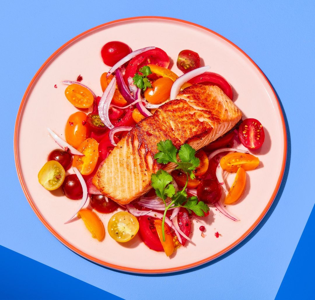 Grilled salmon fillet atop a bed of sliced tomatoes and red onions, garnished with fresh cilantro, presented on a pink plate against a blue background.