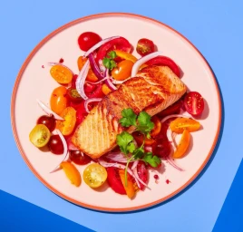 Pan-seared salmon fillet served on a bed of colourful cherry tomatoes and sliced red onion on a pink plate.