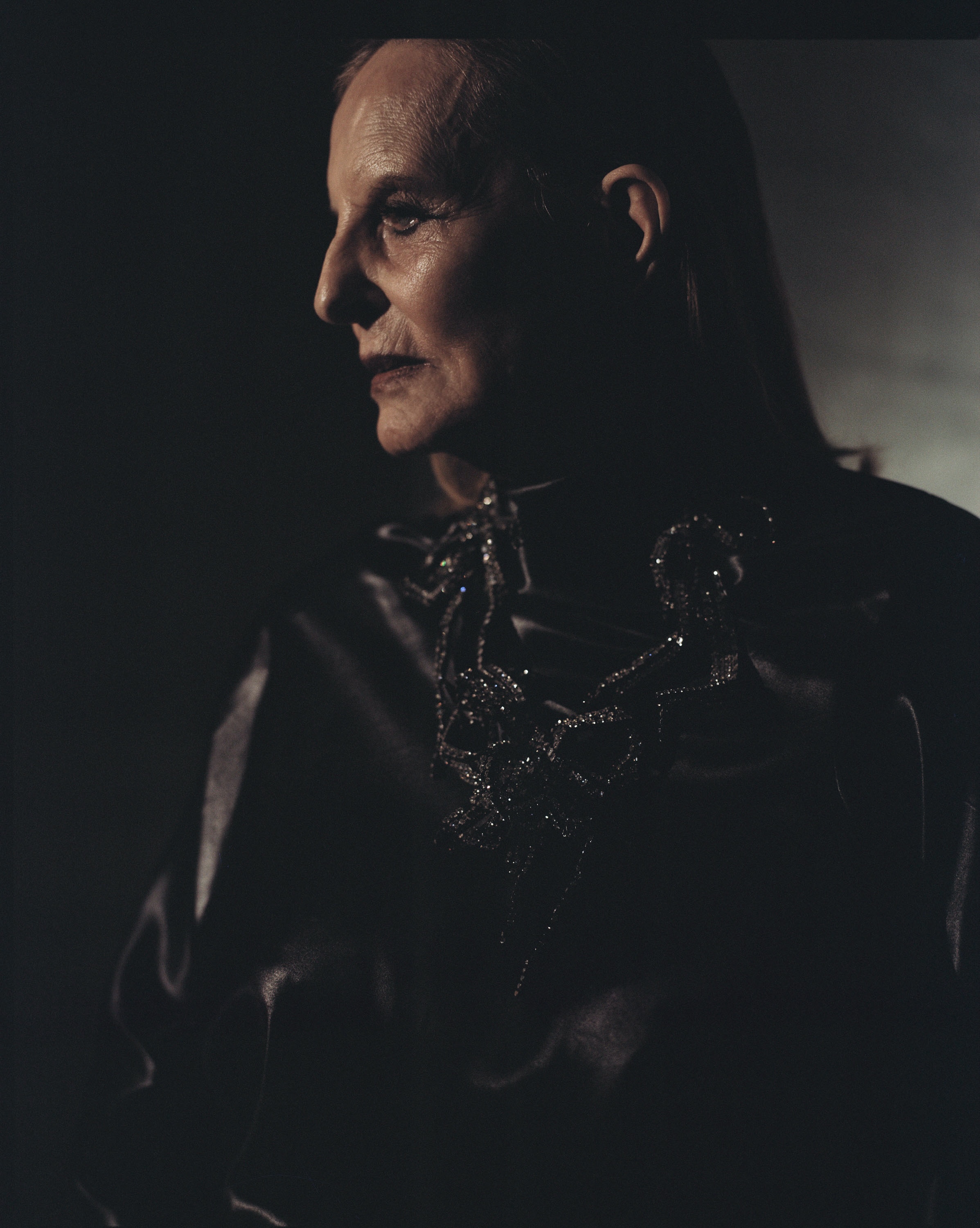 Dramatic studio portrait of an adult with makeup wearing an embellished necklace in dark lighting against a dark background.