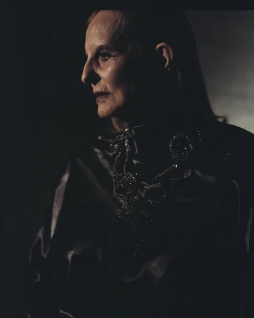 Dramatic studio portrait of an adult with makeup wearing an embellished necklace in dark lighting against a dark background.