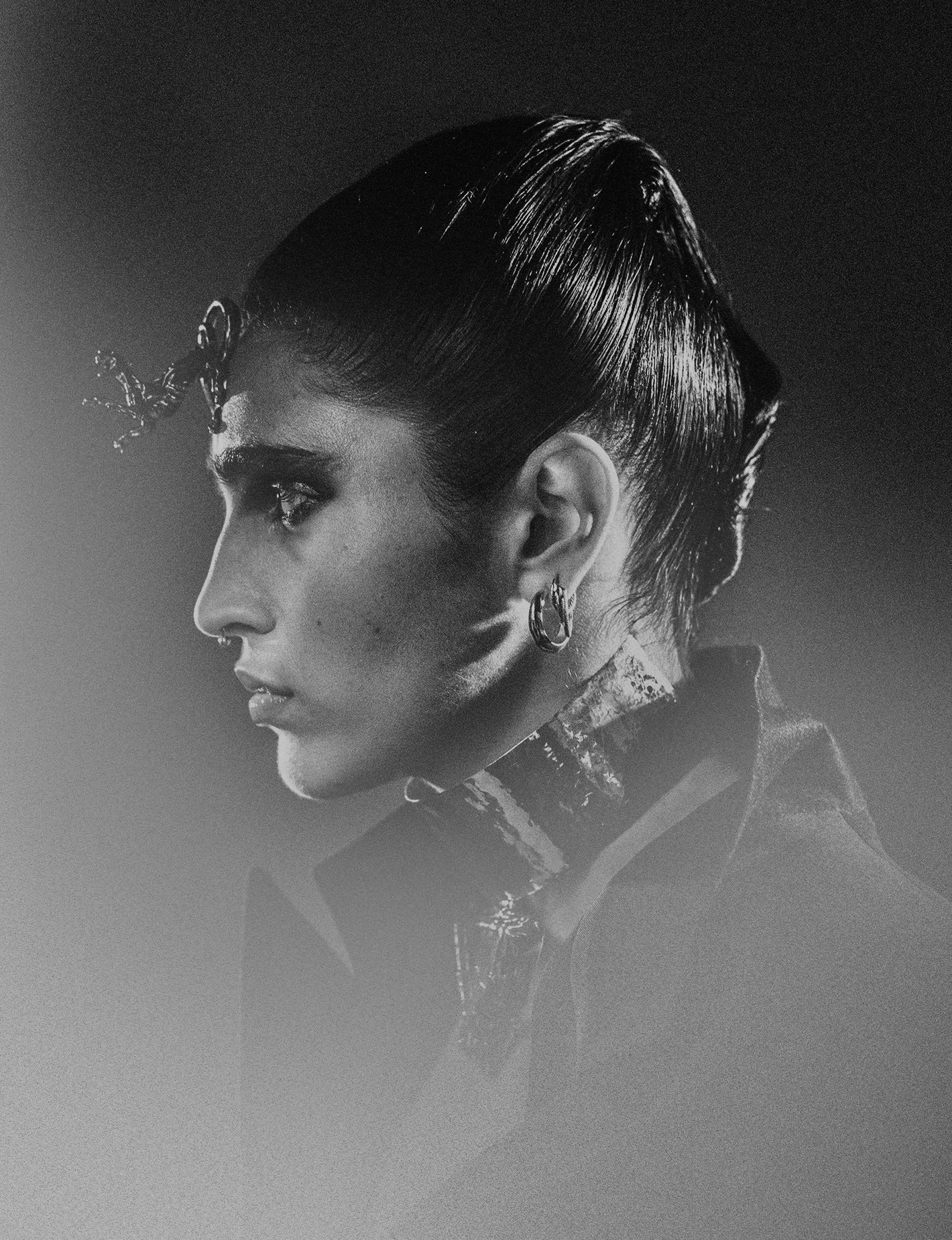 Monochrome portrait of a stylish adult female model in dark clothing and dramatic makeup, featuring distinctive jewelry and slicked-back hair.