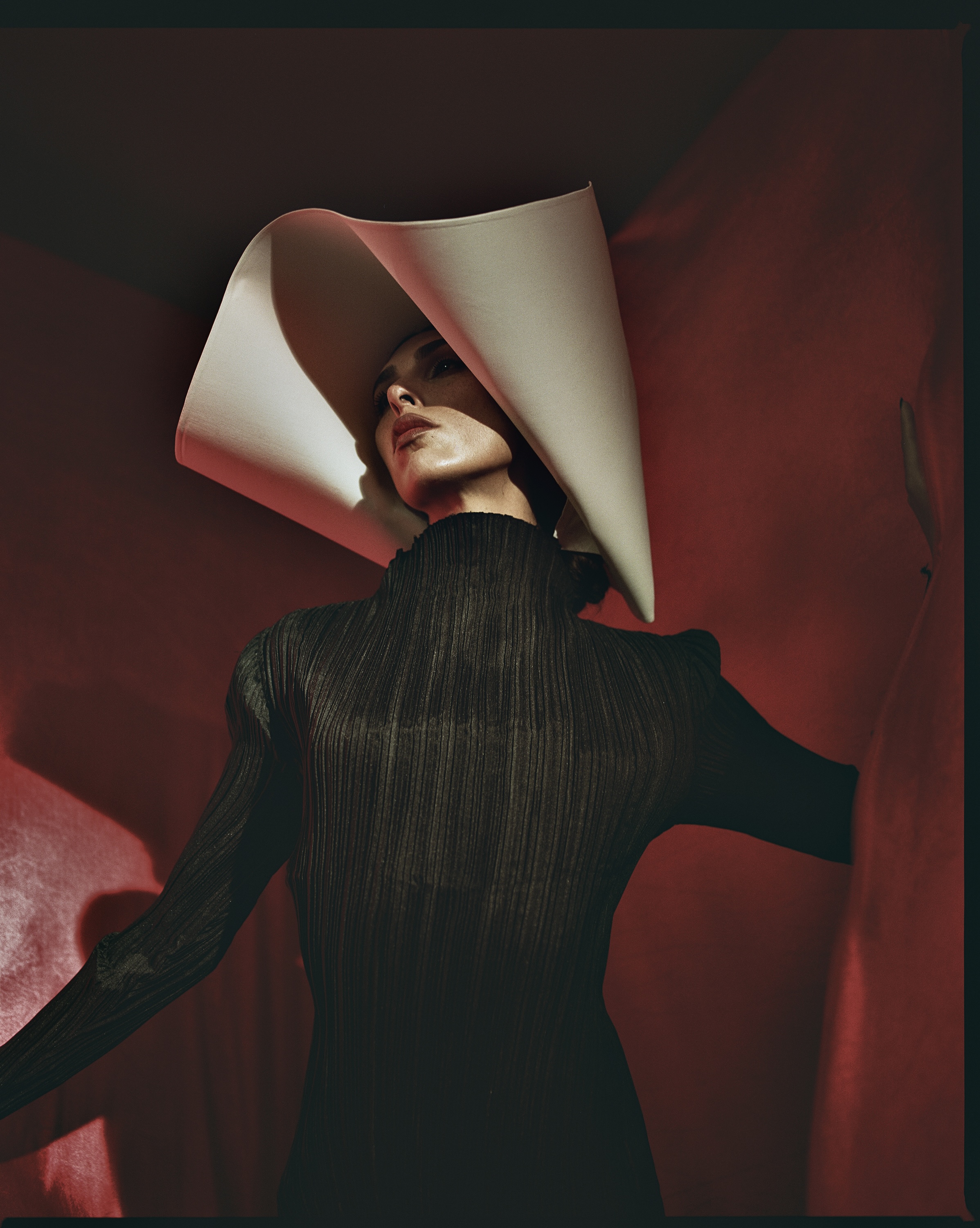 Woman in sculptural white hat and black dress poses against a red backdrop in a studio setting, emphasizing fashion and artistic design.